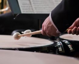 Timpanista di orchestra ripreso di lato dietro una fila di timpani, con le mazze sollevate sopra la pelle