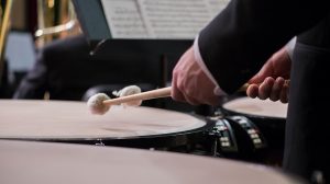 Timpanista di orchestra ripreso di lato dietro una fila di timpani, con le mazze sollevate sopra la pelle