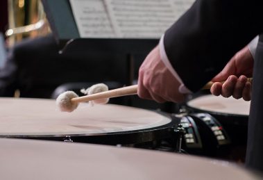 Timpanista di orchestra ripreso di lato dietro una fila di timpani, con le mazze sollevate sopra la pelle
