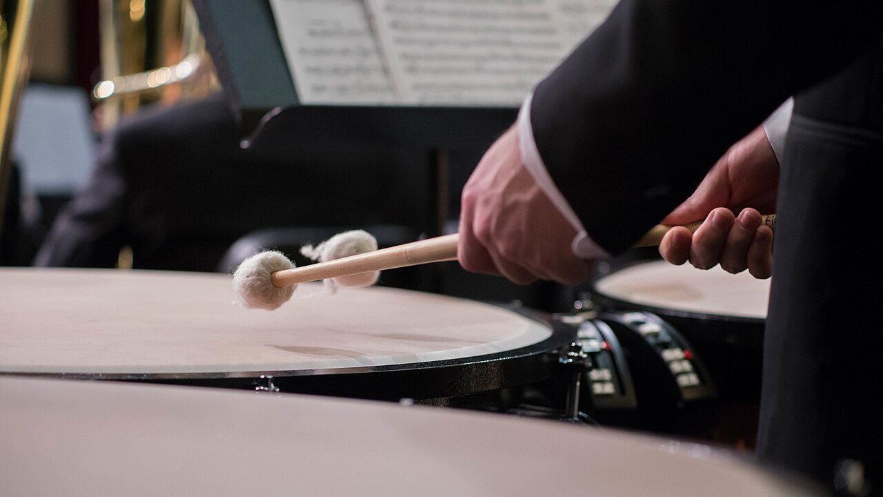 Timpanista di orchestra ripreso di lato dietro una fila di timpani, con le mazze sollevate sopra la pelle