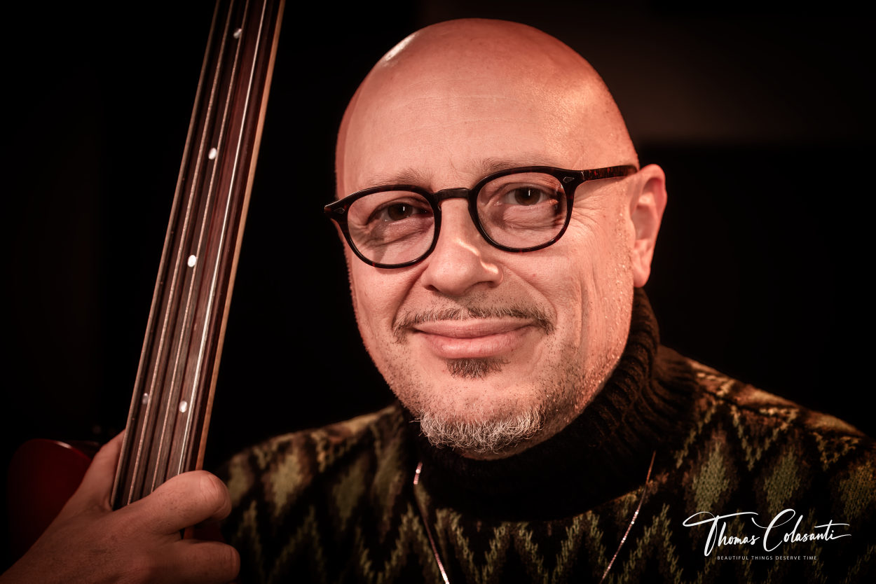 Dario Deidda in studio con il basso rosso di lato, cappellino e occhiali, sorriso disteso