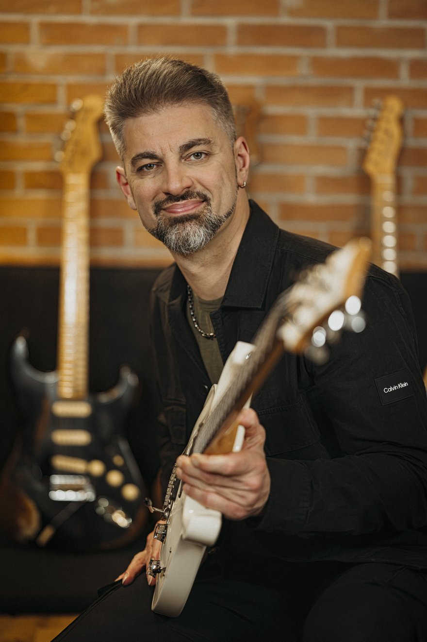 Ciro Manna sorridente con la chitarra elettrica in mano, altre chitarre visibili sullo sfondo dello studio