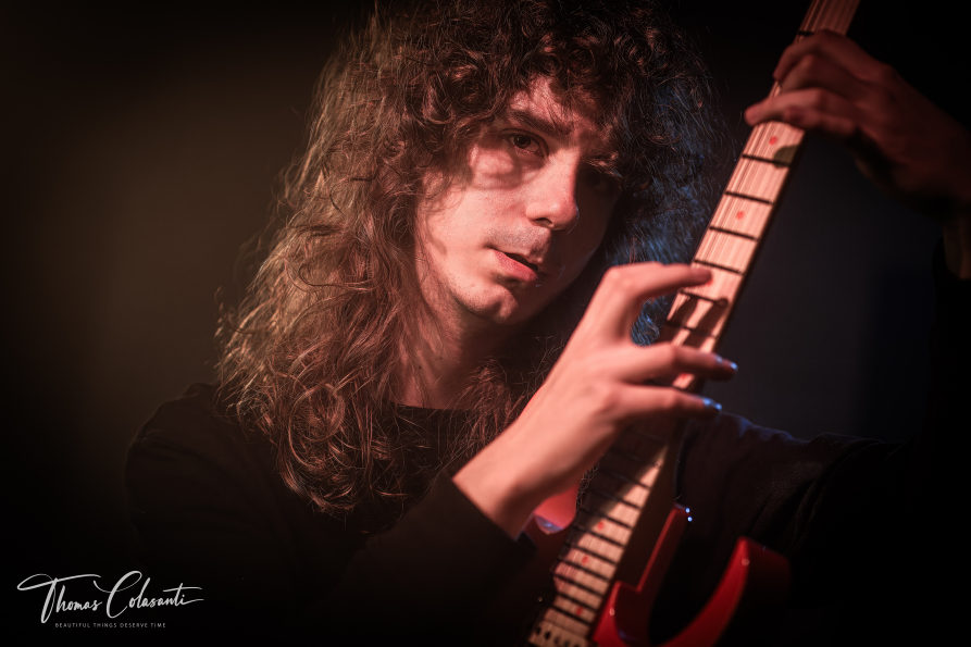 Edoardo Taddei in primo piano estremo, guancia appoggiata al manico della chitarra elettrica rossa mentre la mano sinistra preme sui tasti, luce blu di scena sullo sfondo.