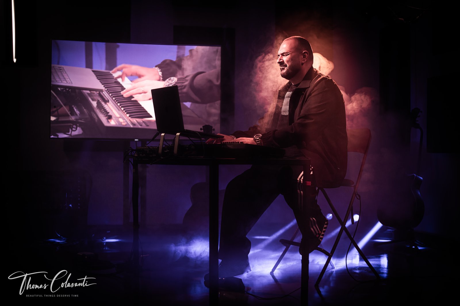 Max D'Ambra seduto alla consolle del suo studio con laptop e tastiera controller, uno schermo alle spalle proietta un primo piano di mani sui tasti, fumo di scena e luci blu e viola