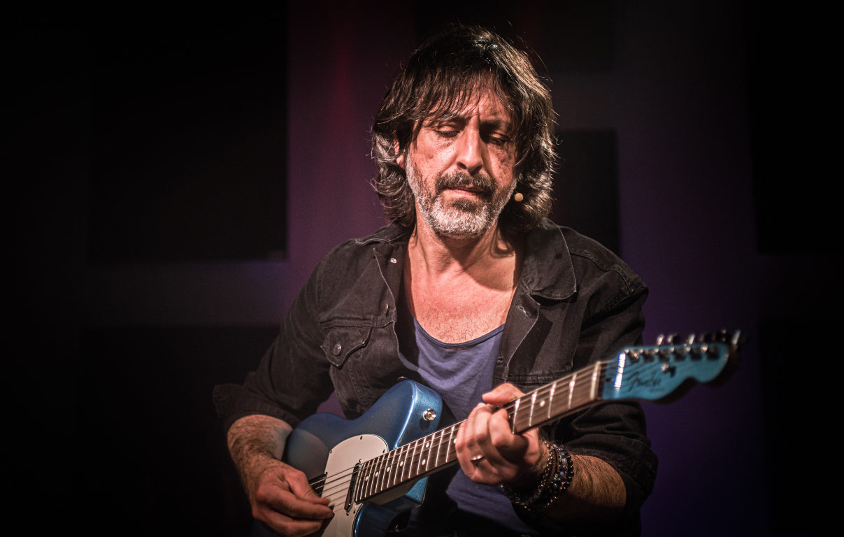 Osvaldo Lo Iacono seduto in ripresa media con la chitarra elettrica blu ben visibile, sfondo con luci viola e rosse, sguardo abbassato sul manico