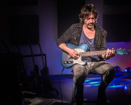 Osvaldo Lo Iacono seduto su uno sgabello con una chitarra elettrica blu, sguardo abbassato verso lo strumento, rack di chitarre sullo sfondo