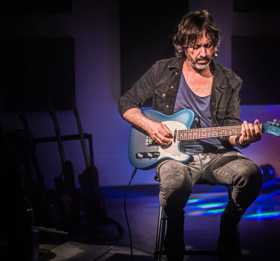 Osvaldo Lo Iacono seduto su uno sgabello con una chitarra elettrica blu, sguardo abbassato verso lo strumento, rack di chitarre sullo sfondo