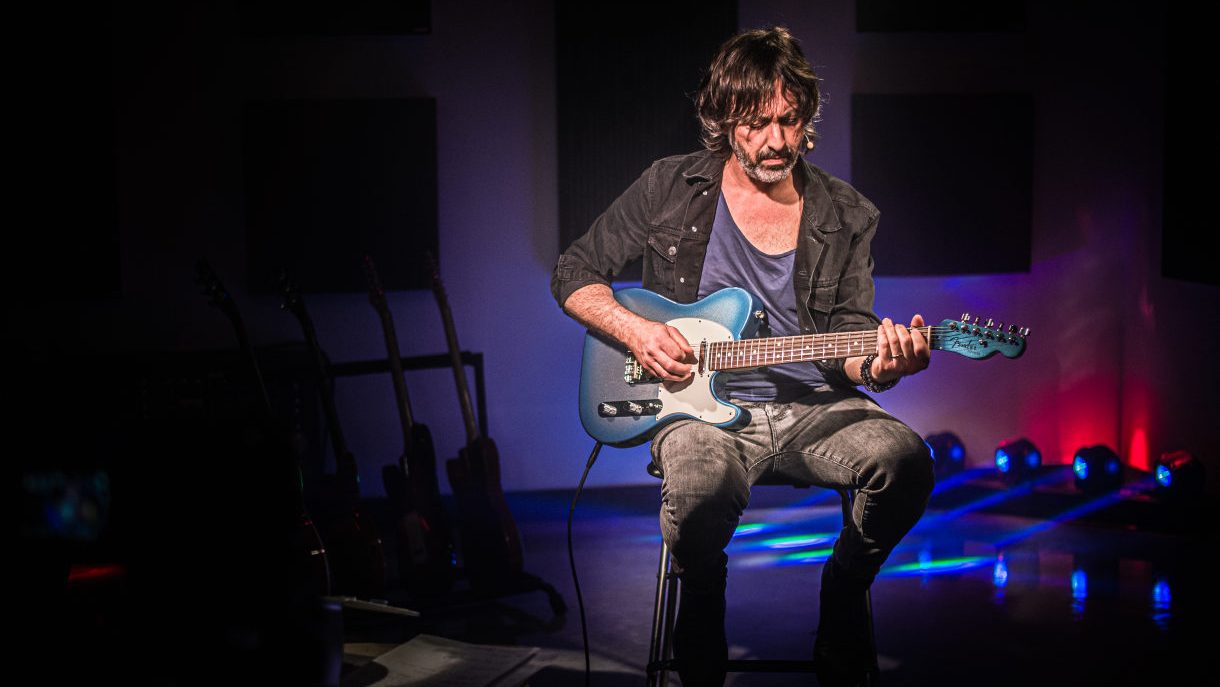 Osvaldo Lo Iacono seduto su uno sgabello con una chitarra elettrica blu, sguardo abbassato verso lo strumento, rack di chitarre sullo sfondo