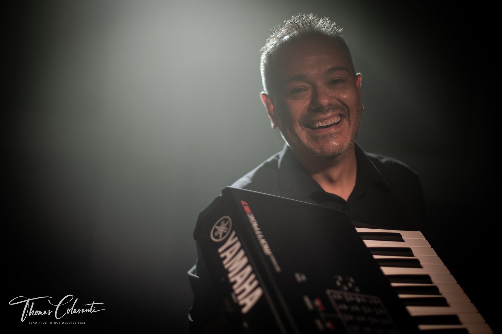 Manuele Montesanti sorride in controluce mentre solleva una tastiera Yamaha, luce di scena verdastra alle spalle