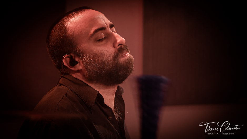 Gabriele Morcavallo ripreso di profilo con occhi chiusi ed espressione rilassata, kit batteria sfocato sullo sfondo, luce rossa calda da studio
