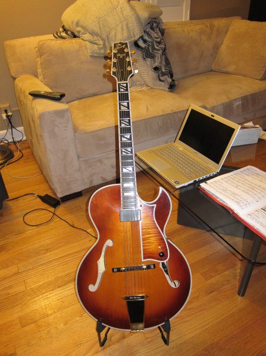 Una chitarra archtop Heritage Sweet 16 con finitura sunburst appoggiata a uno stand, vista di fronte con cordiera, ponte in legno e tastiera intarsiata
